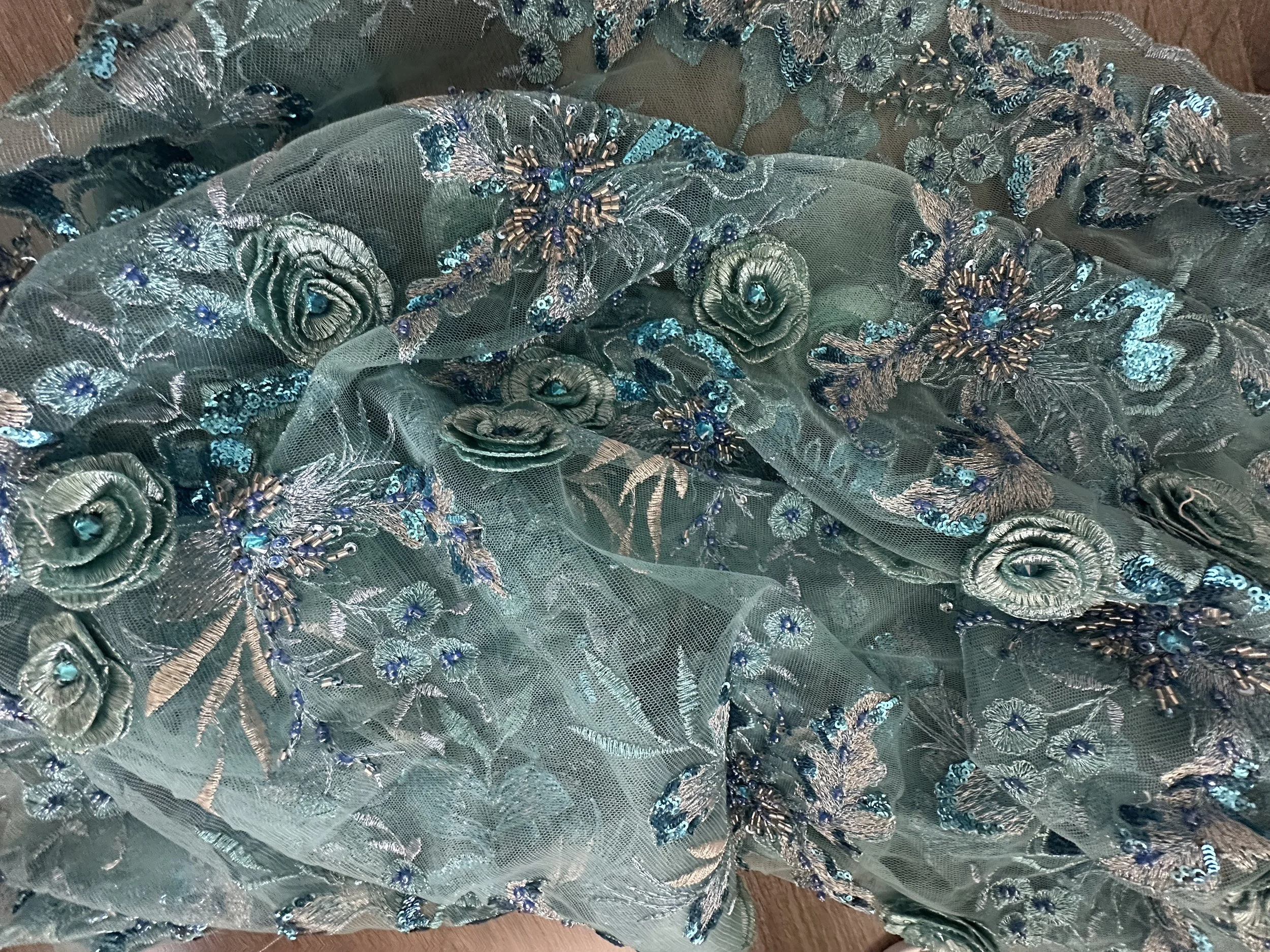 Close-up of a teal-colored fabric with intricate floral embroidery, featuring roses, leaves, and sparkly embellishments, possibly part of a gown or decorative textile.