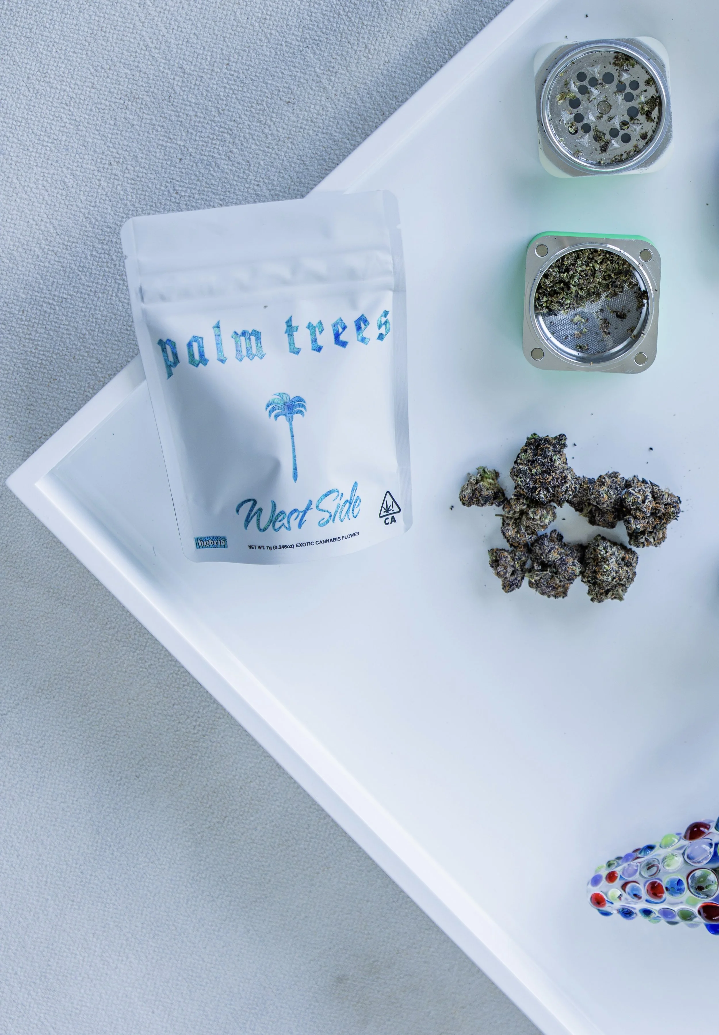 A white tray with cannabis buds, a bag labeled 'palm trees West Side', two circular metal screens with cannabis, and a glass marble filled with colorful swirls.