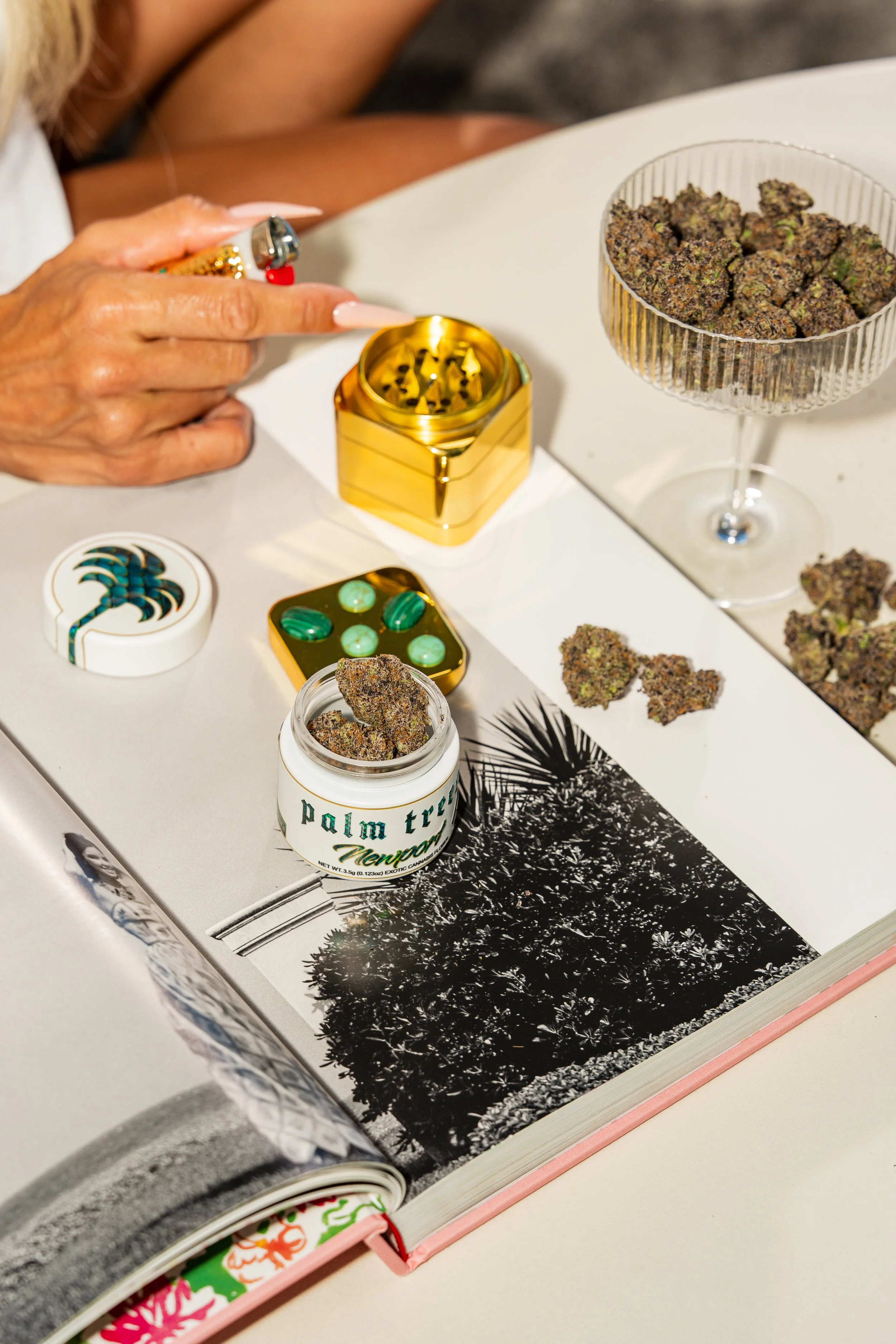 A person holding a lit lighter with cannabis buds on a white table. There are containers of cannabis and a book with black and white trees on the pages.