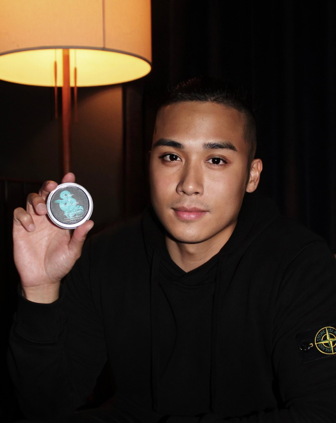 A young man with short black hair and a black hoodie holding a round badge with a blue duck design.