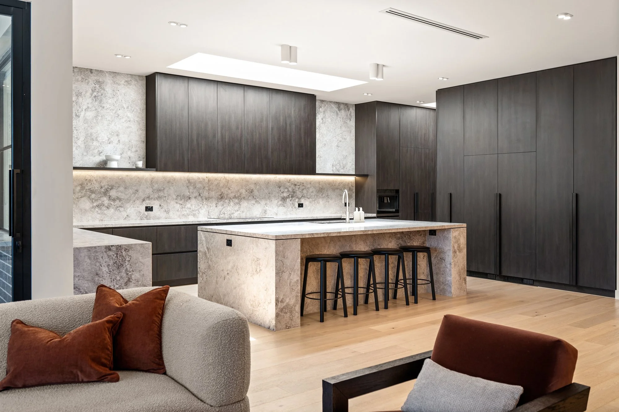 Modern kitchen with dark wood cabinets, a marble island with four black stools, and a marble backsplash with recessed lighting.