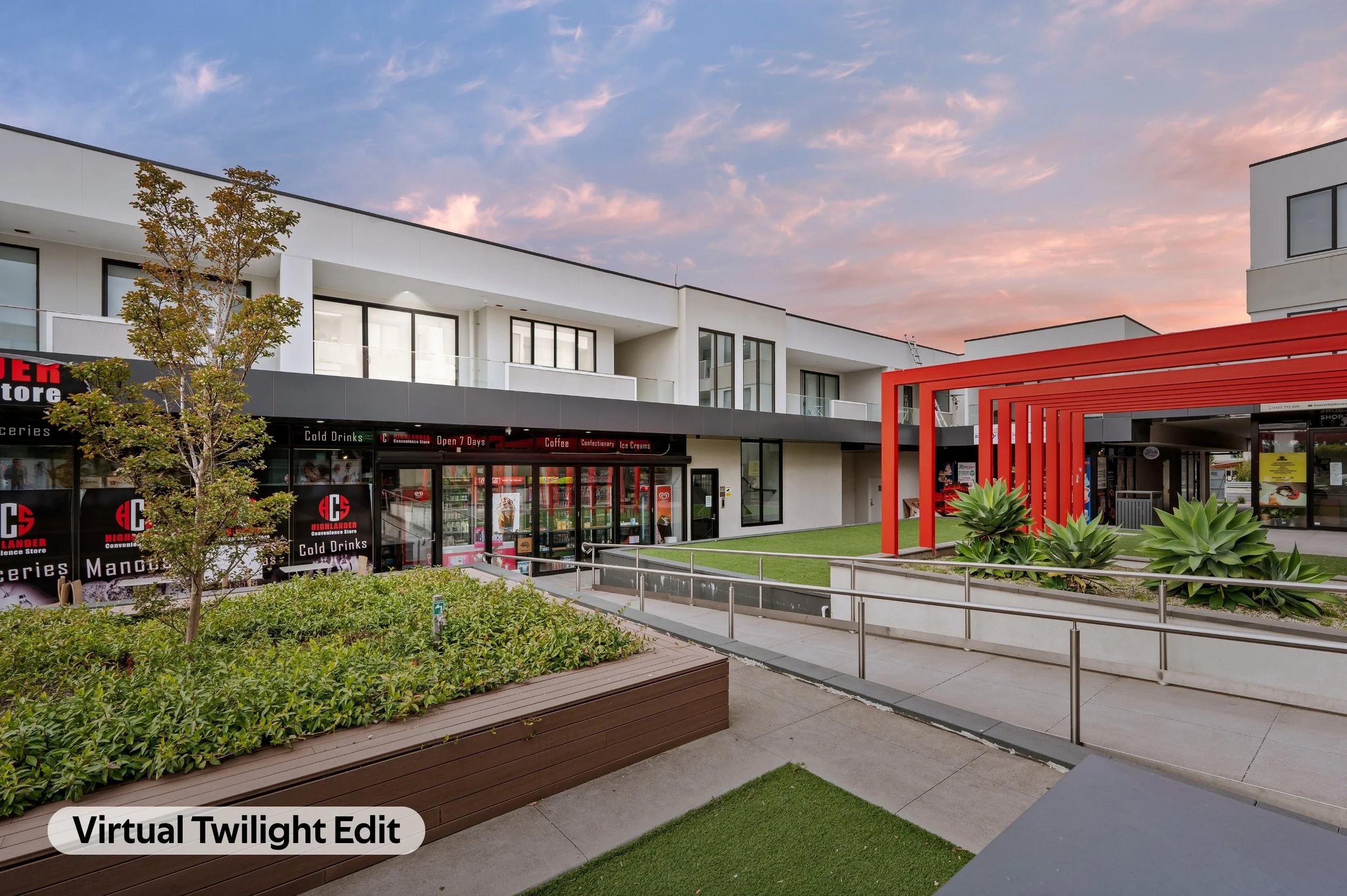 A modern commercial or residential complex featuring white buildings with black-framed windows, a small tree with green and brown leaves, a landscaped area with green bushes and succulents, a red architectural structure, and a sky with pink and blue 