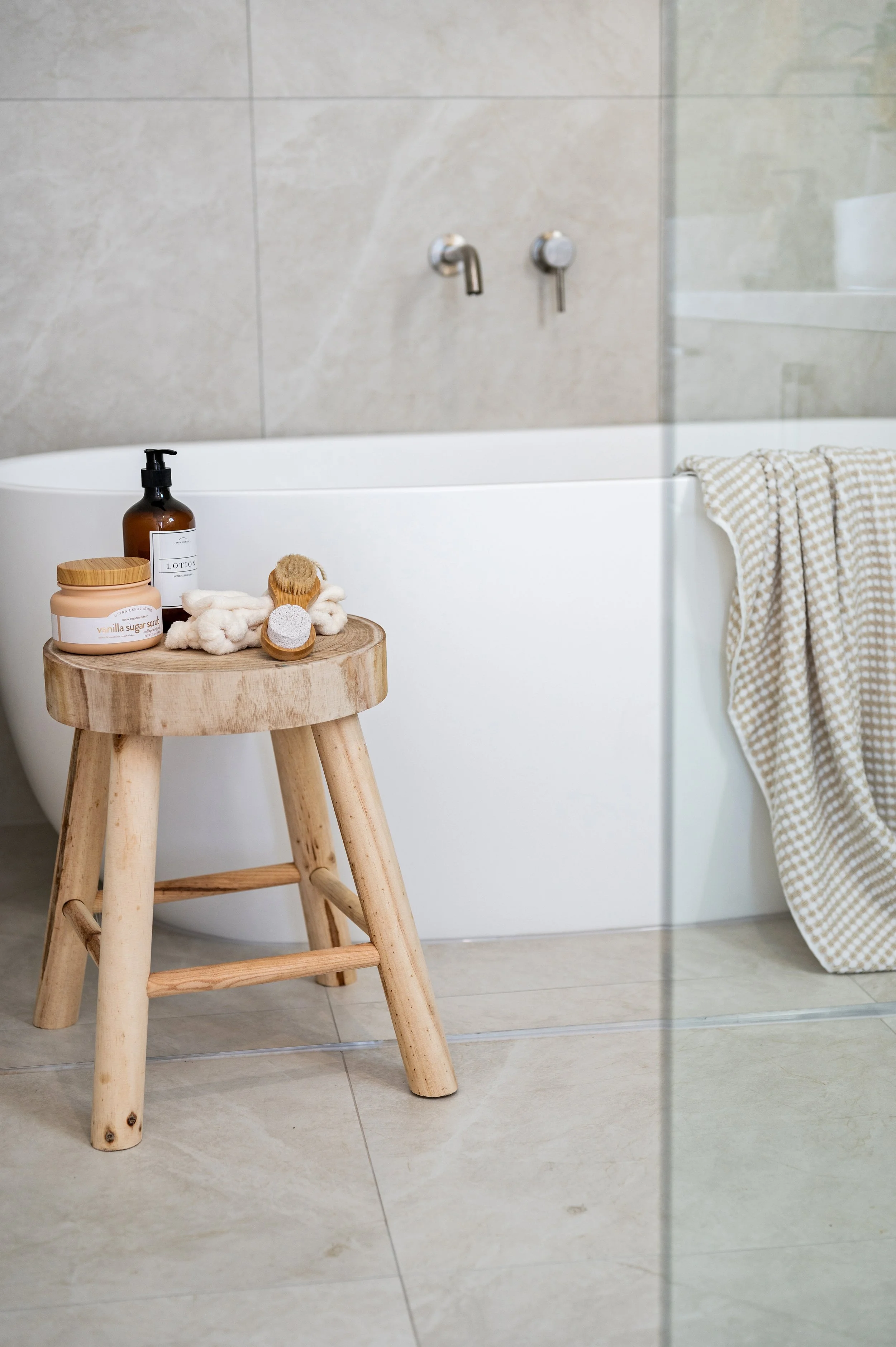 Top of a round wooden stool with toiletries on it, including a soap dish, lotion bottle, and scrub, next to a bathtub with a towel hanging on the side, in a bathroom with tile walls and floor.