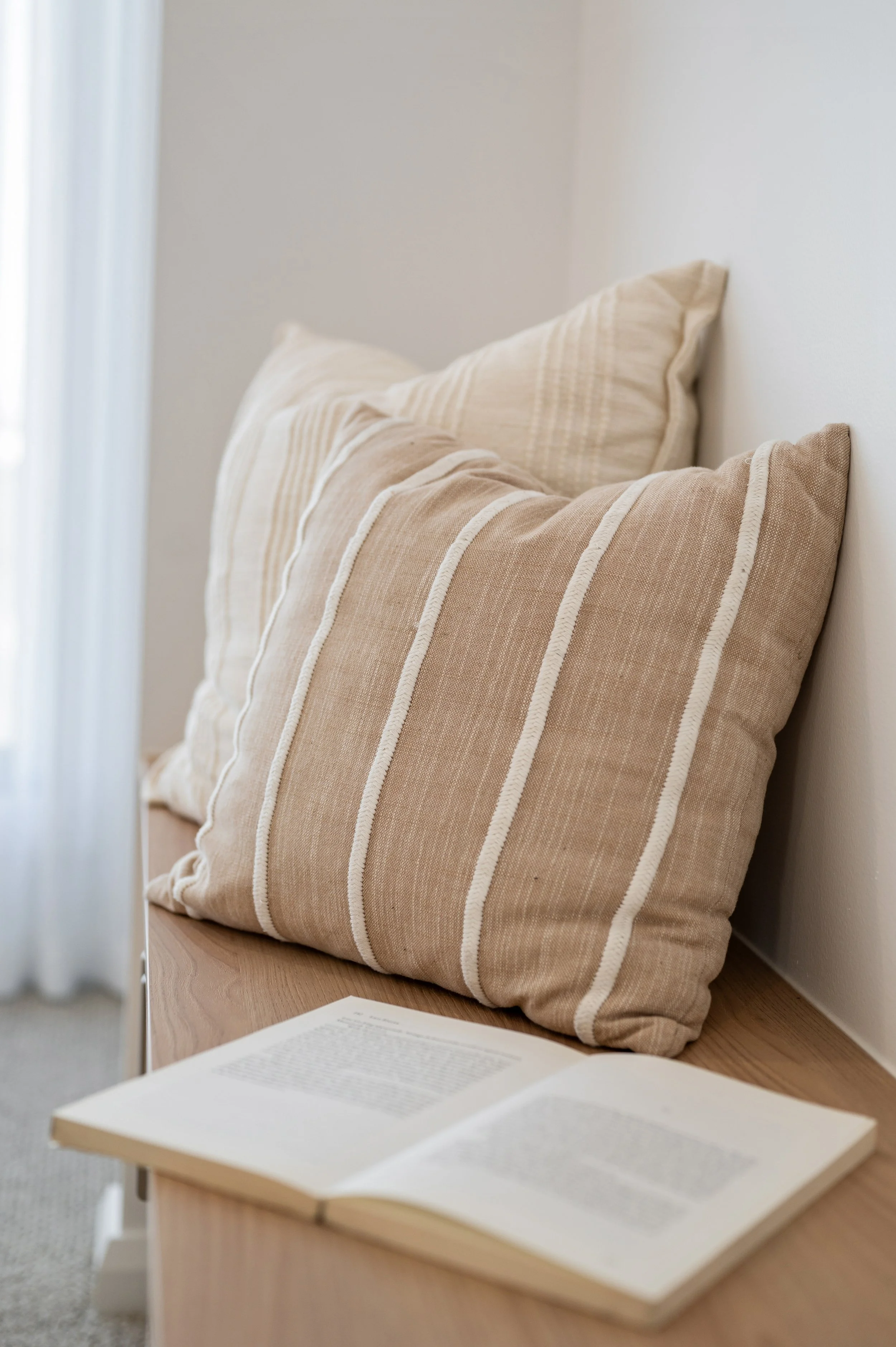 Two beige striped pillows on a wooden bench with an open book in the foreground, against a light-colored wall.