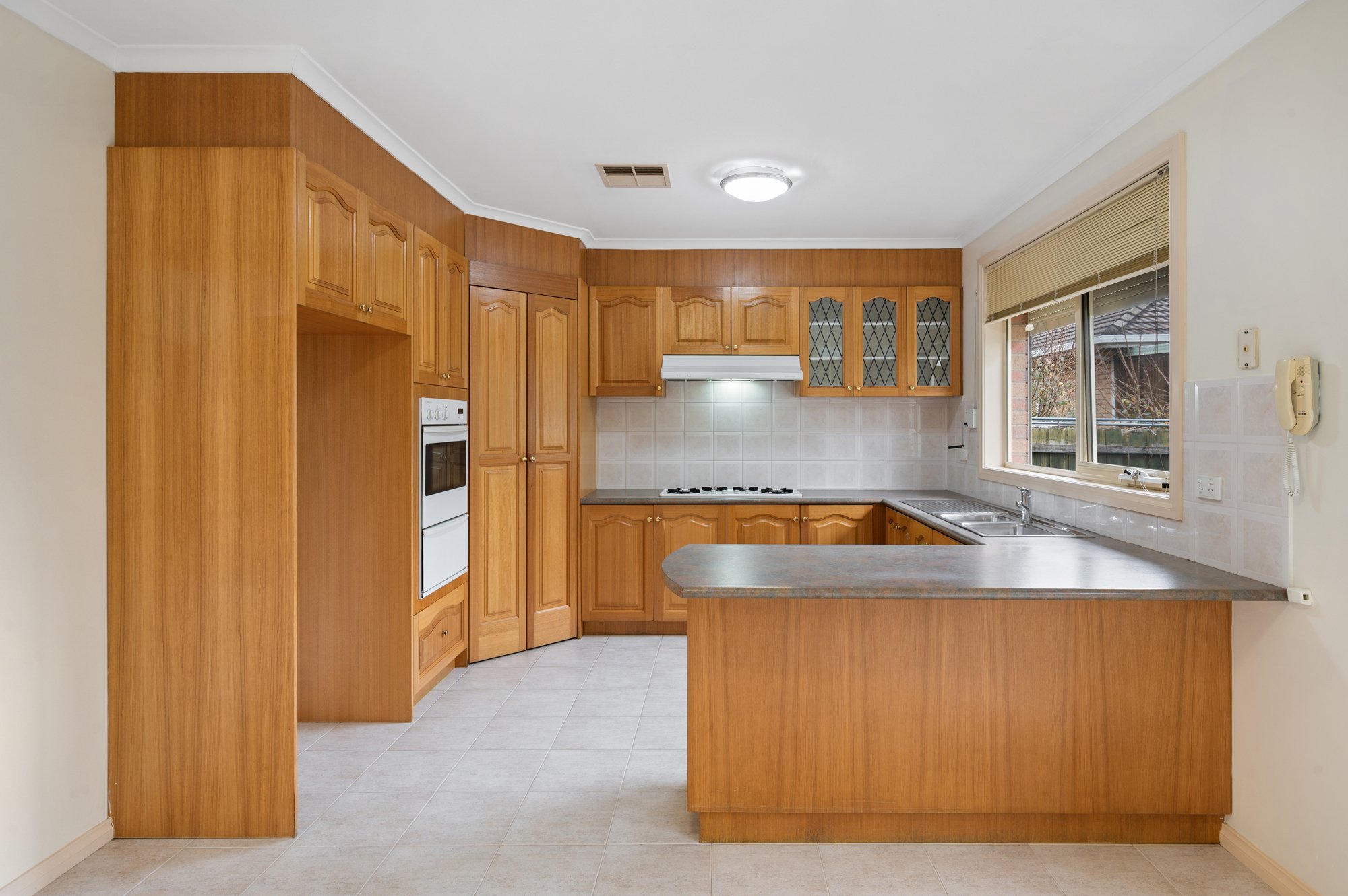 Kitchen with wooden cabinets, beige tiled backsplash, window with blinds, and a white oven above a microwave.