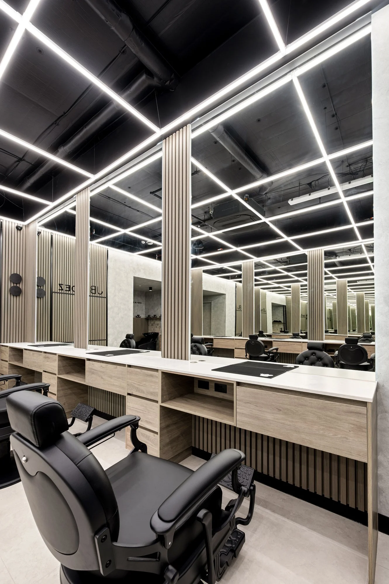 Modern salon or barbershop with black chairs, light wood stations, and a ceiling with illuminated grid pattern, featuring black ducts and sleek lighting.