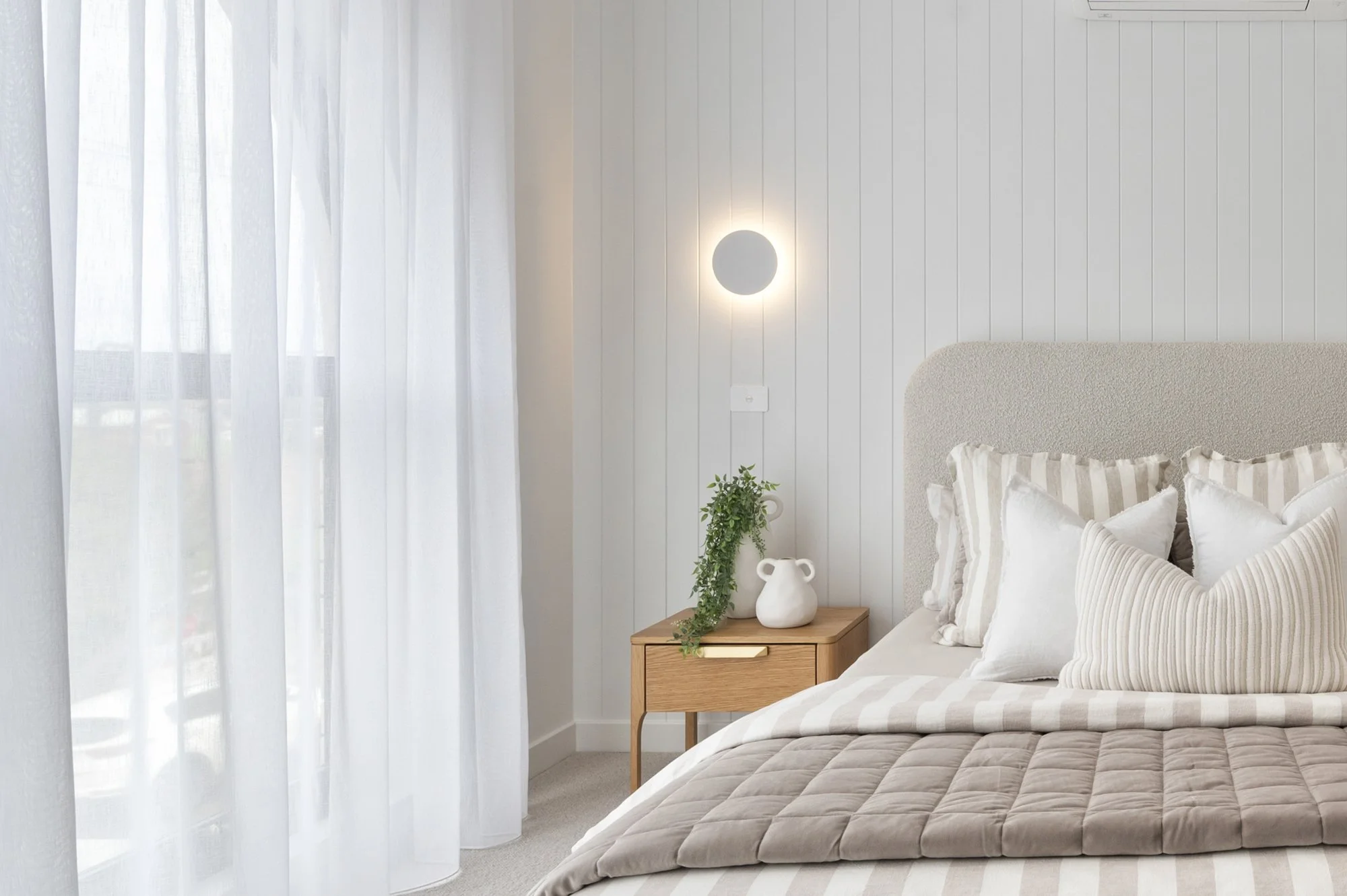 Bright bedroom with sheer white curtains, a textured beige bedspread, and a small wooden nightstand with a potted green plant and a white decorative vase. There is a white wall with vertical paneling and a round wall light above the nightstand.