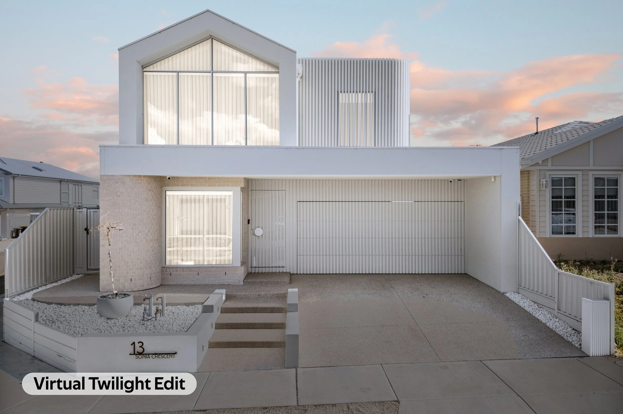 Modern white house with a balcony, vertical slat design on the upper level, a large front window with curtains, and a small garden area with white gravel and a potted plant in the front yard.