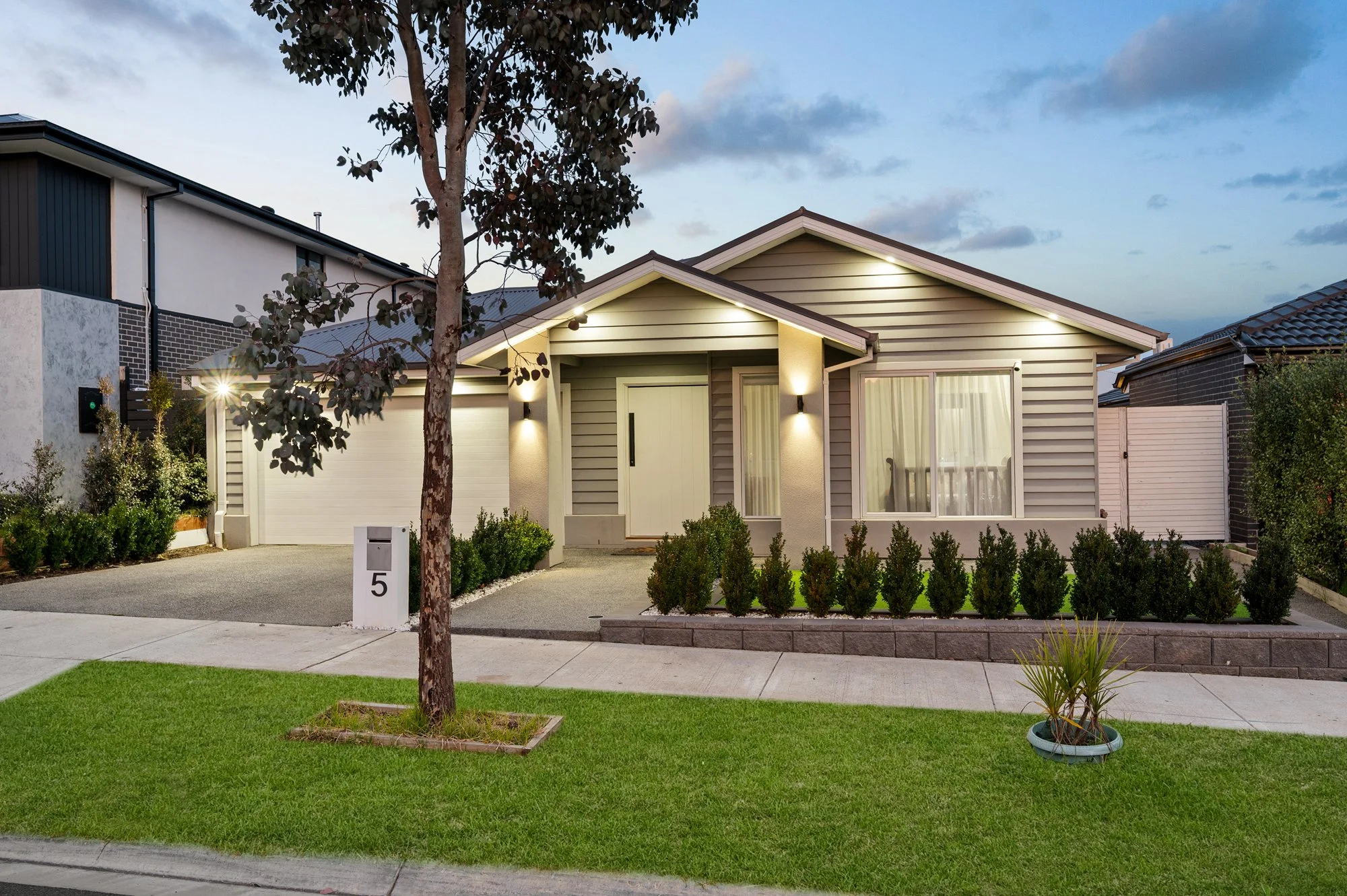 Front view of a modern single-story house with gray horizontal siding, a white front door, and large front windows. The house is illuminated with outdoor lights and has a well-maintained green lawn with small bushes and a potted plant. There is a tree in the foreground and neighboring buildings on either side. The sky is partly cloudy at dusk.