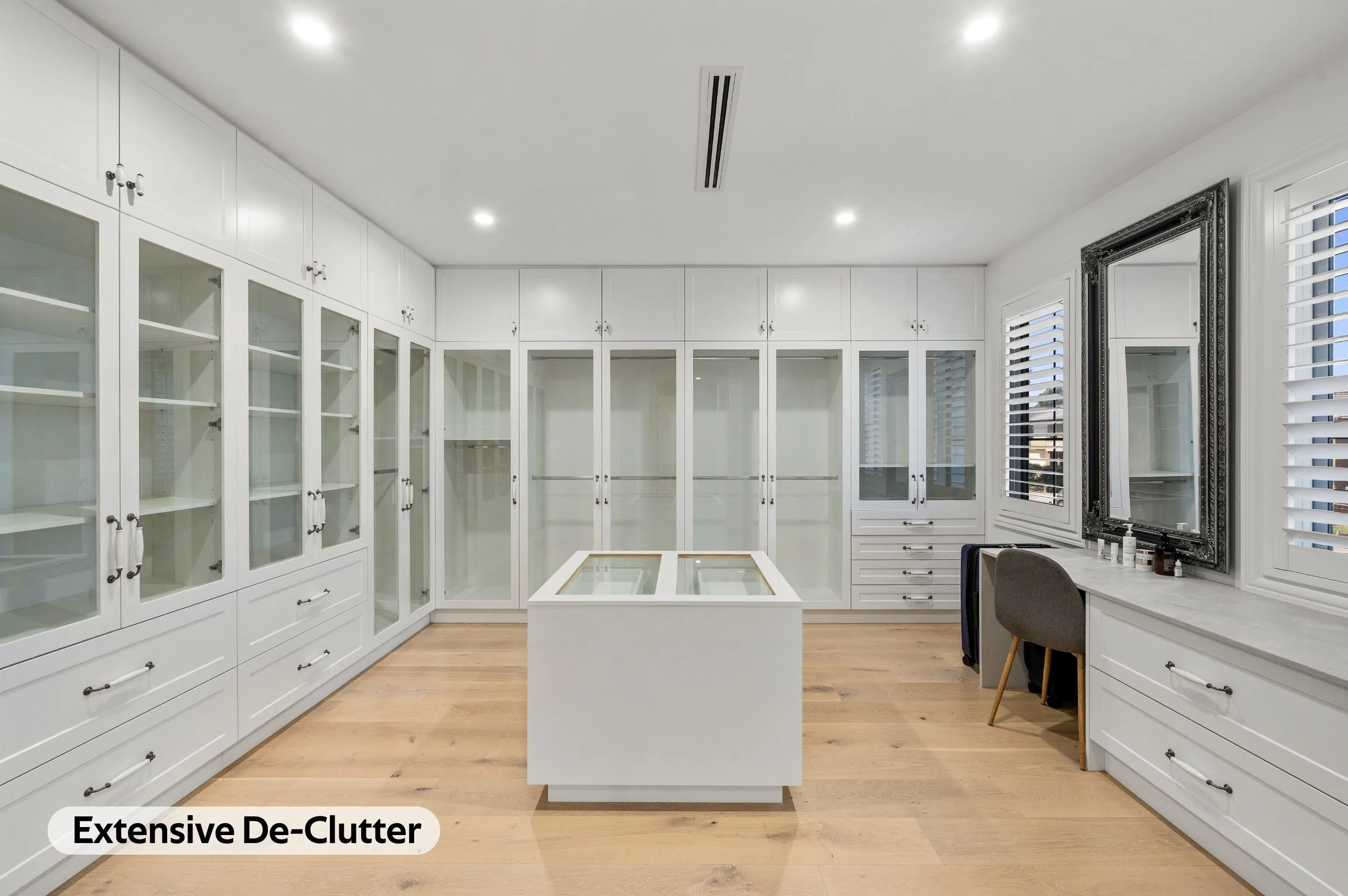 A spacious walk-in closet with white cabinets and glass-fronted shelves, a large window with plantation shutters, a decorative mirror, and light hardwood floors.