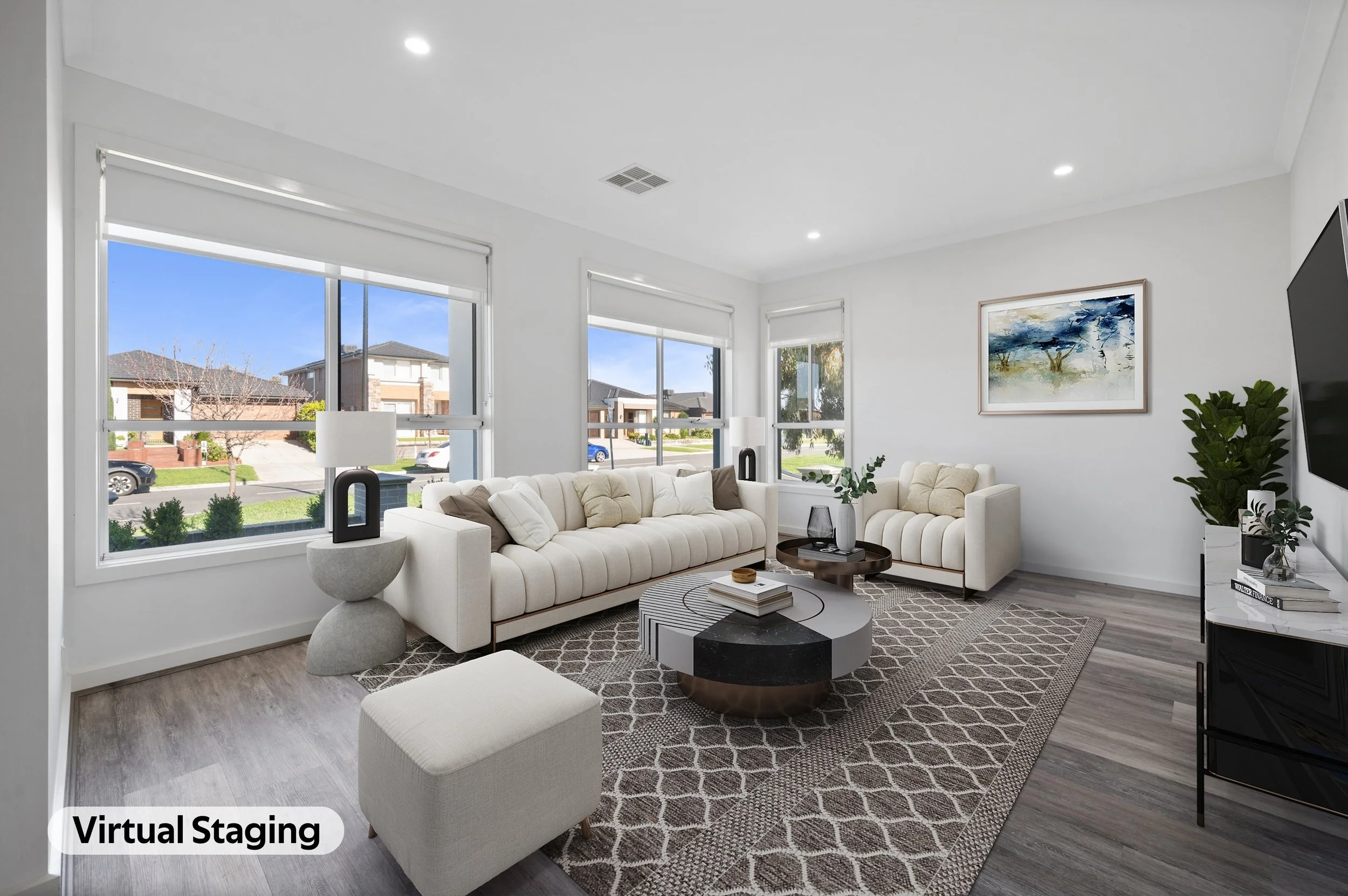 Living room with white sofas, a coffee table, large windows showing a suburban neighborhood, artwork on the wall, a potted plant, and a patterned rug.