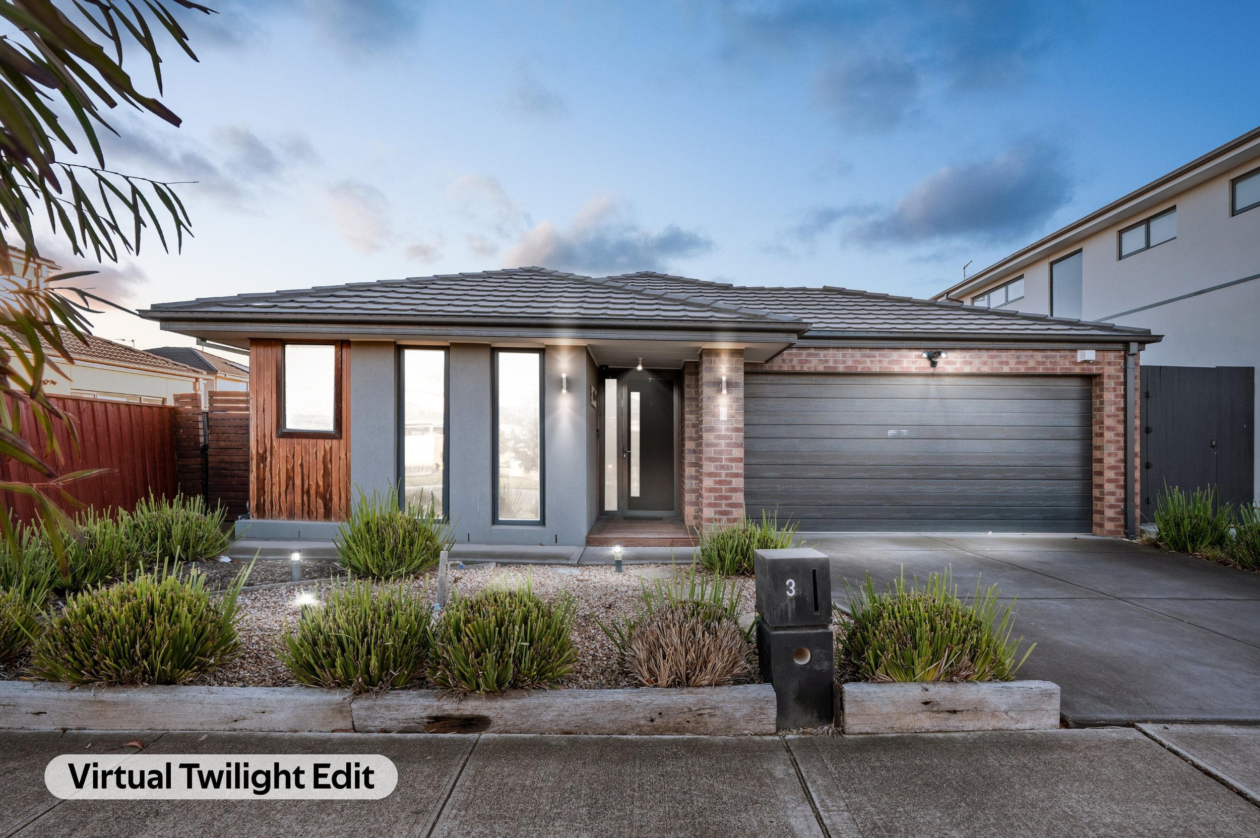 Modern house with a gray garage door, brick accents, and front entrance illuminated by exterior lights. Landscaping includes low shrubs and a gravel bed, with a wooden border and pathway leading to the door. The sky is partly cloudy with a twilight a