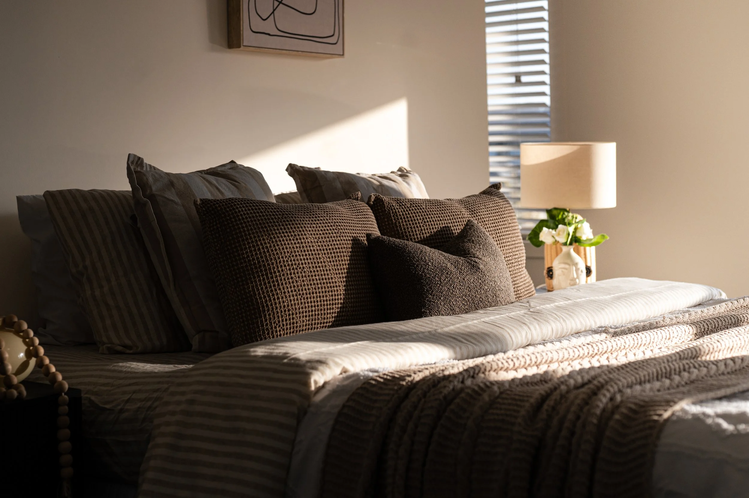 A neatly made bed with multiple pillows in front of a plain wall. To the right, there is a bedside lamp and a small vase with white flowers. Sunlight is streaming through a window with horizontal blinds, casting light and shadows.