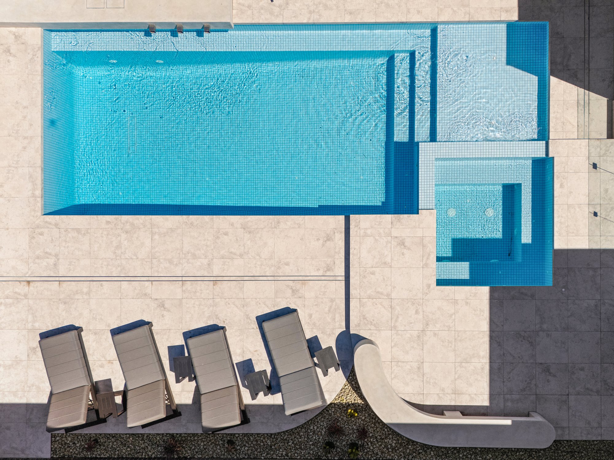 Top-down view of a swimming pool with a tiled interior, surrounded by beige paving stones. Six lounge chairs are lined up to the left of the pool, casting bright shadows.