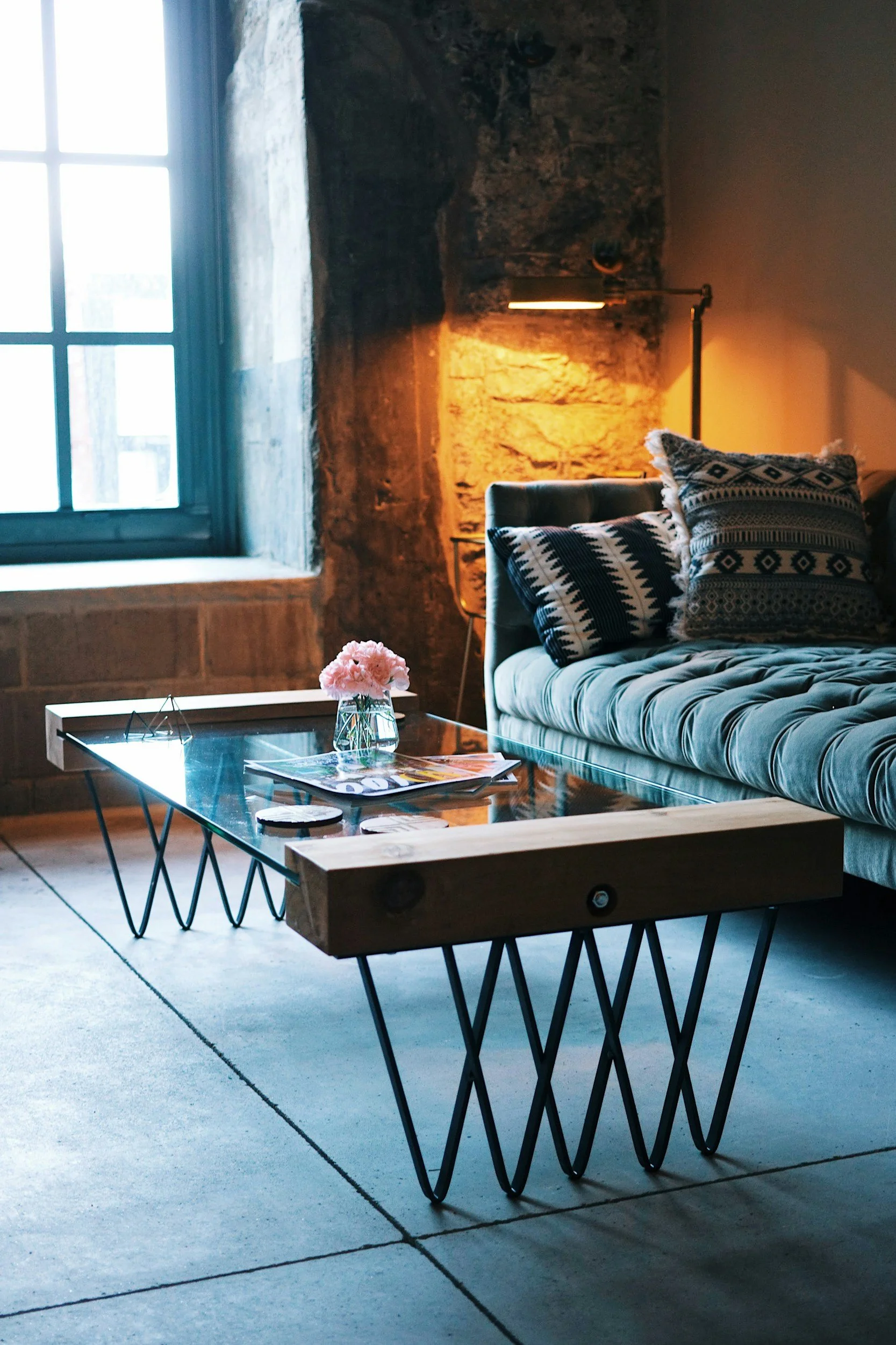 Cozy living room with a large window, stone wall, mustard-colored lamp, patterned pillows on a plush sofa, glass-top coffee table with pink flowers, magazines, and small decorative items, on a concrete floor.