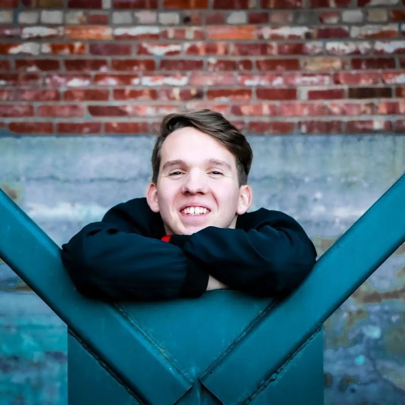 A young man with short brown hair leaning on a teal-colored metal structure, smiling with his arms crossed, in front of a brick wall backdrop.