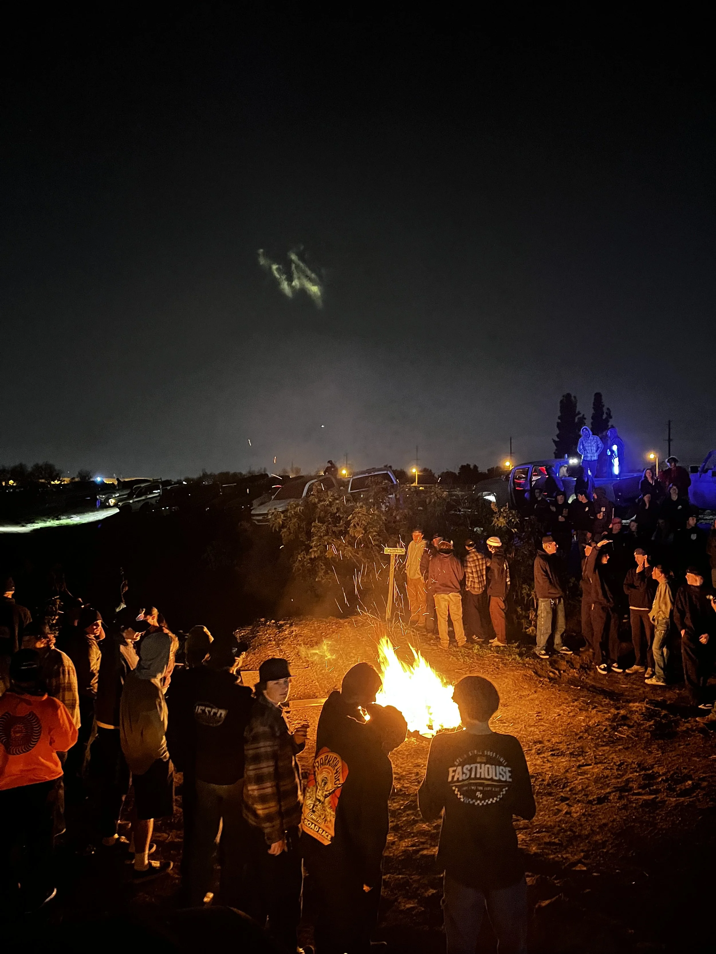 Group of people gathered around a campfire at night with parked cars and trees in the background.