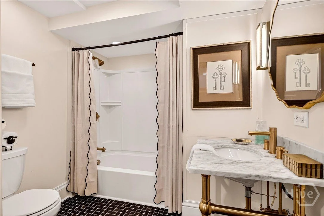 Bathroom with a bathtub and shower, white toilet with a sheep figurine on top, beige curtains, framed artwork, marble vanity with a gold faucet, and black tile flooring.
