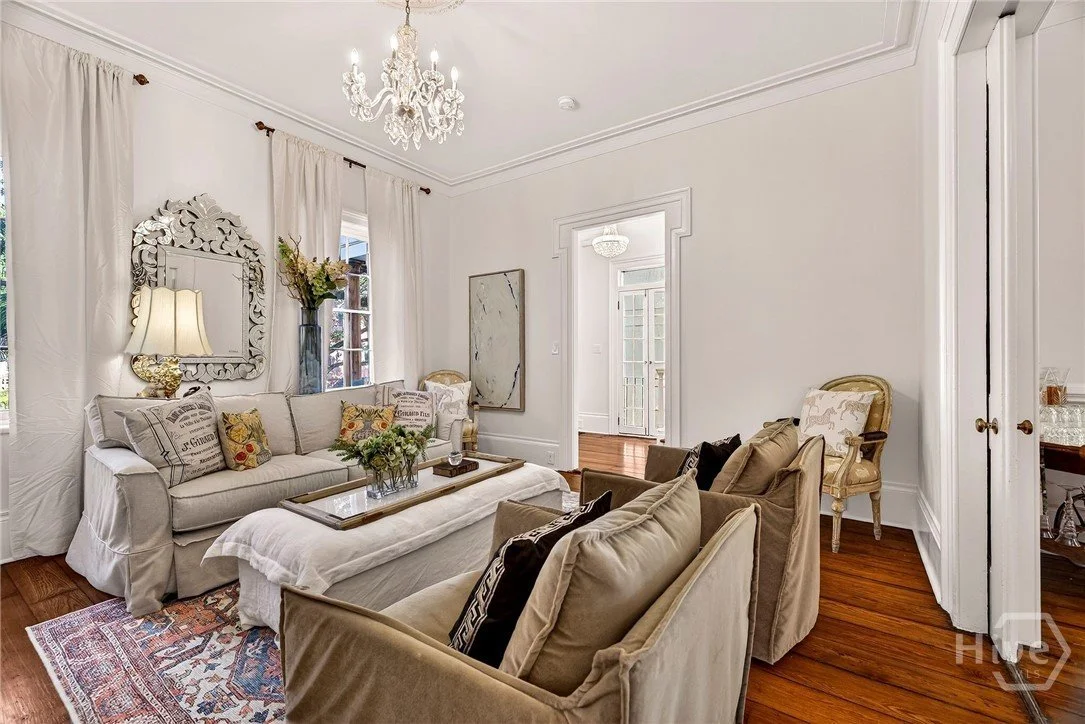 Living room with white walls, a chandelier, a sofa with multiple pillows, armchairs, a coffee table with a flower arrangement, a mirror, and windows with white curtains.