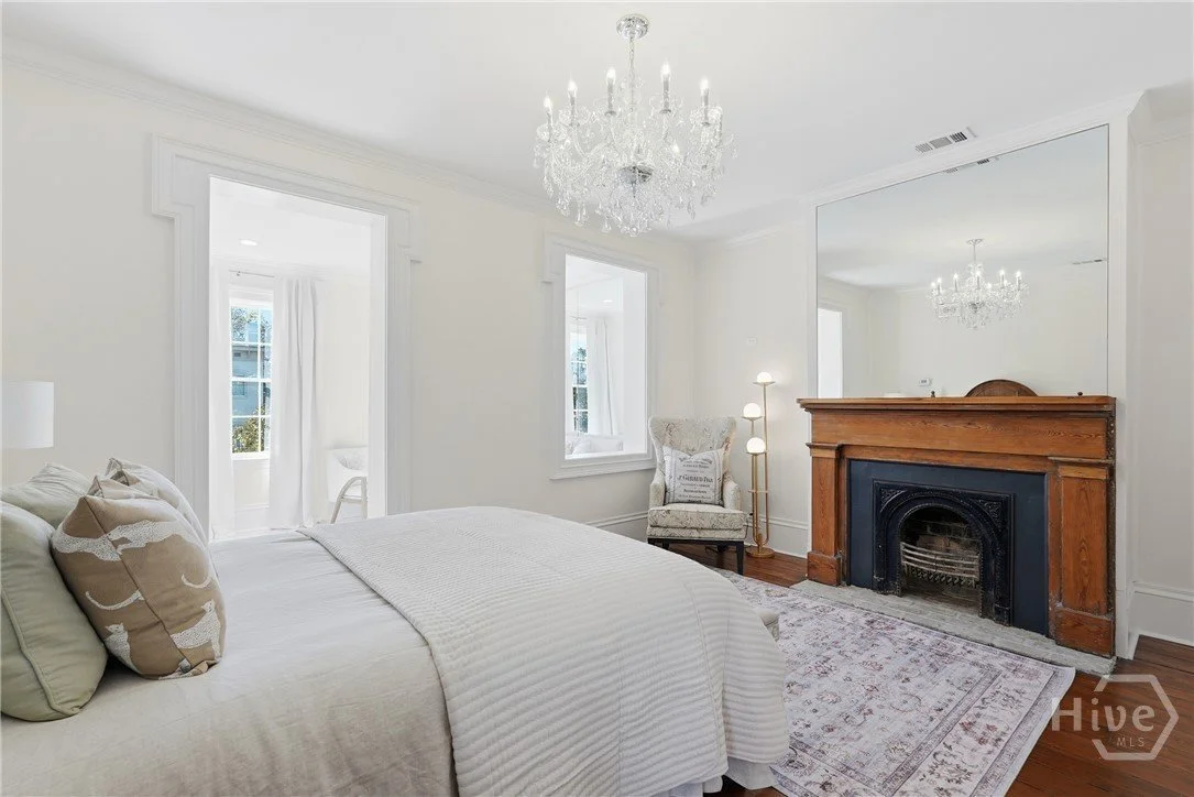 A bedroom with a white bed, decorative pillows, a chandelier hanging from the ceiling, a fireplace with a wooden mantle, a floor lamp, and an upholstered armchair near the window.