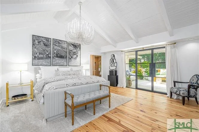 Bright bedroom with white walls, large bed, artwork of zebras above, chandelier, two lamps, a bench, and a sliding glass door leading to a balcony with greenery outside.