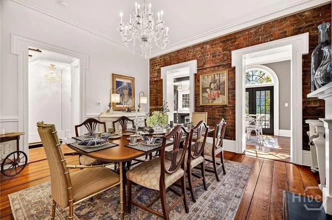 A dining room with a wooden table, ten chairs, a white fireplace, artwork on the wall, a chandelier, and an exposed brick wall.