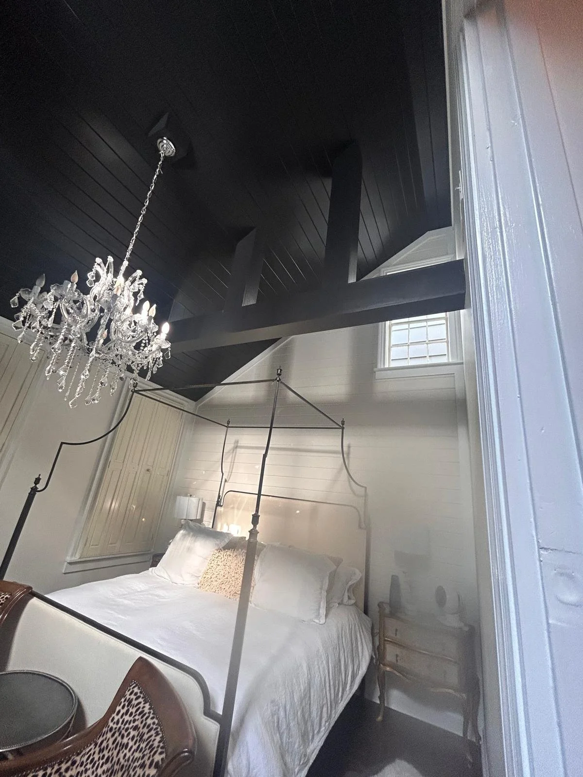 A bedroom with a four-poster bed with white bedding and a decorative pillow, nightstands on each side, a chandelier hanging from a dark ceiling, a window above the bed, and a reflection of the room in a mirror.