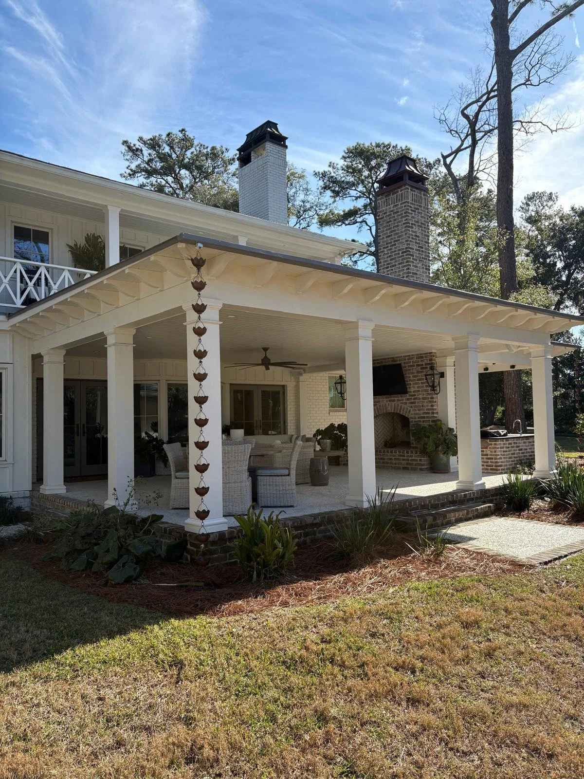 A covered outdoor patio with white columns and woven furniture, facing a garden with green plants, bushes, and a grassy lawn, with a house featuring brick and white siding and a chimney in the background.
