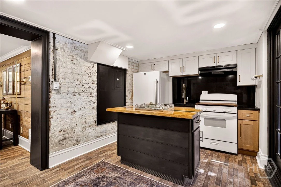 Kitchen with black and white cabinetry, a wooden countertop on the island, a white stove, a white refrigerator, a black backsplash, and a brick wall.