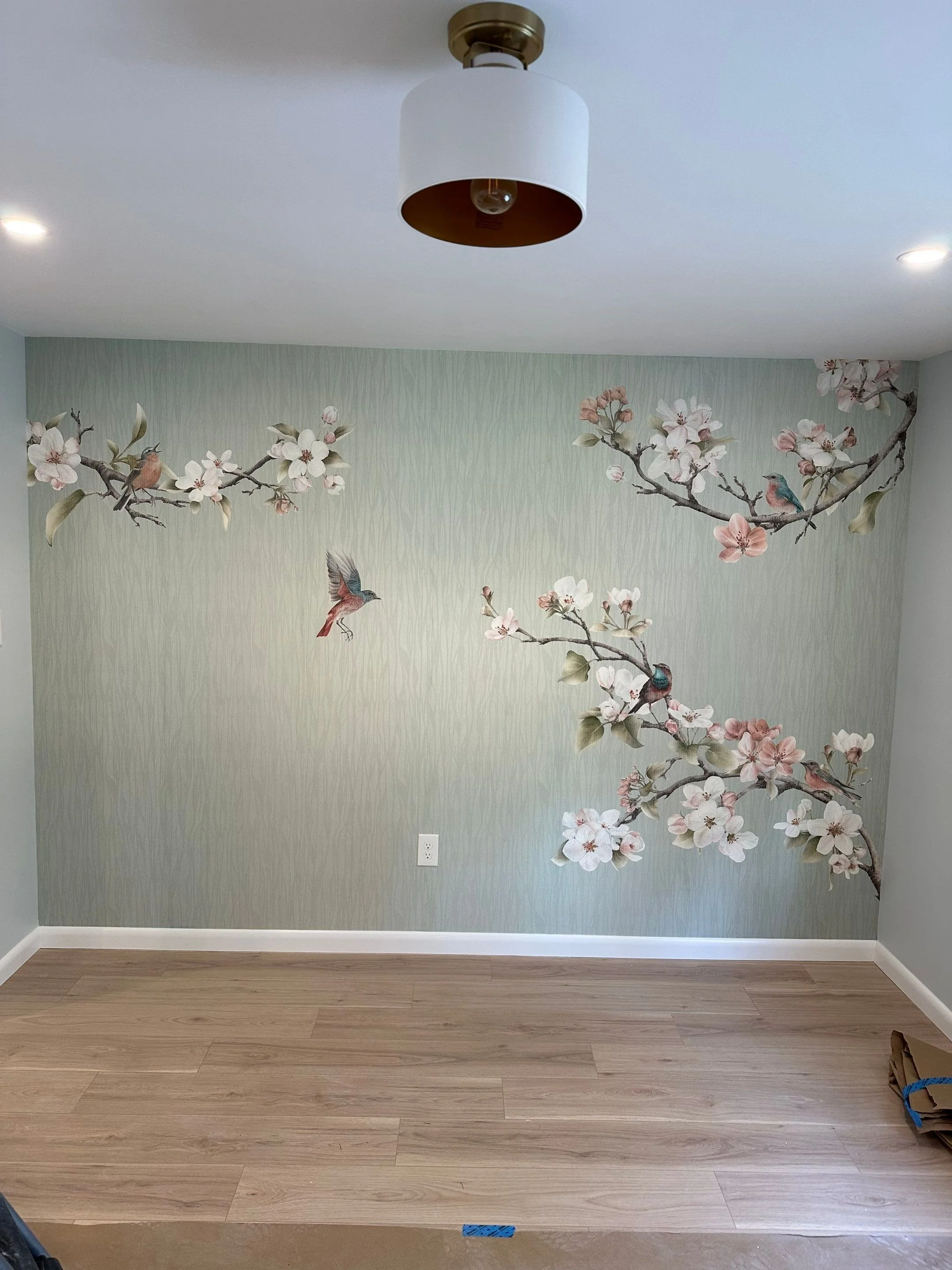 Wall with mural of cherry blossom branches, birds, and a hummingbird on a light green background, with hardwood flooring and ceiling light fixture.