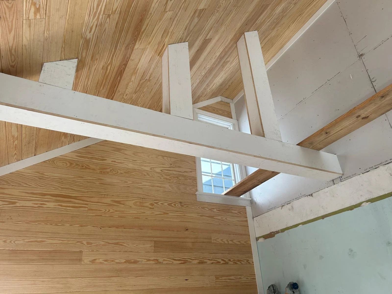 Interior view of a building under construction with wooden flooring, partially constructed white framing, and a window showing a blue sky.