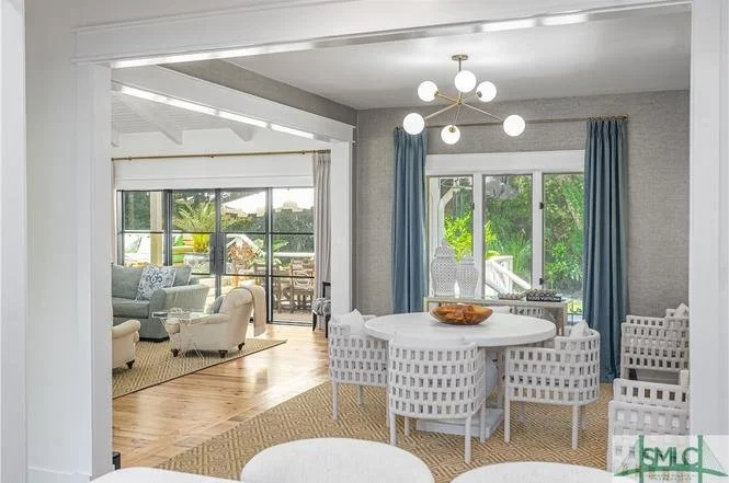 Interior view of a bright living and dining area with large windows, blue curtains, and modern lighting fixture, with outdoor greenery visible through the windows.