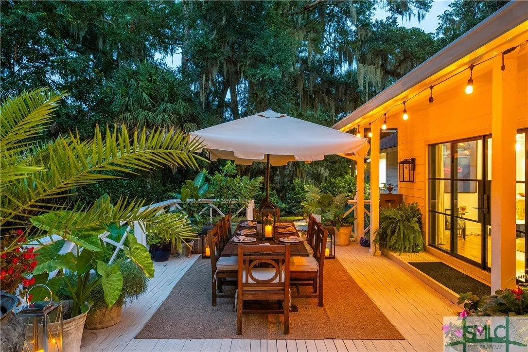 An outdoor dining area on a deck surrounded by lush green plants, with a table set for a meal under a large white umbrella, candle lanterns, and string lights illuminated along the side of a yellow house.
