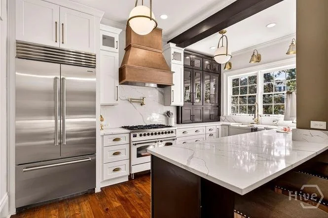 Modern kitchen with white cabinets, a stainless steel refrigerator, a white and gold gas stove with a copper range hood, black upper cabinets, large windows, and a white marble island with dark base.