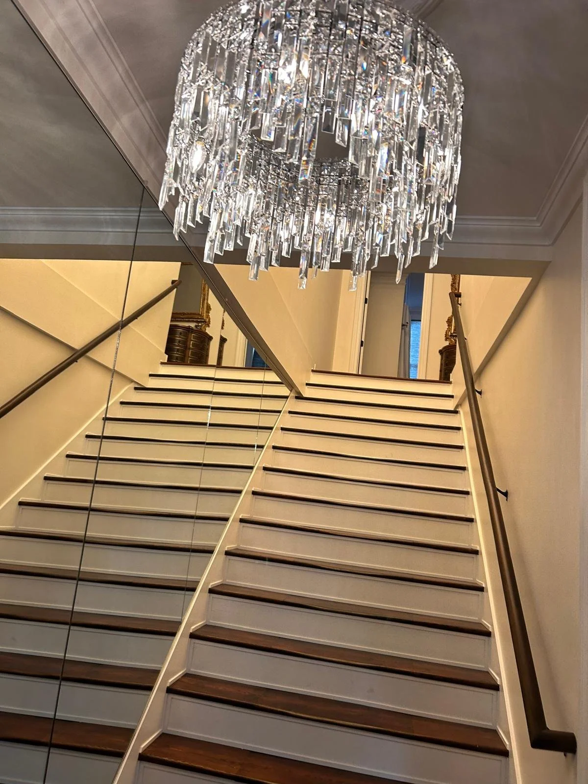 View of a staircase with wooden steps and black handrails, a large crystal chandelier hanging from the ceiling, and a mirror reflecting part of the staircase and chandelier.