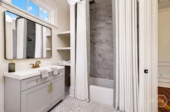 Bathroom with light gray vanity, gold hardware, mirror cabinets, and a single handle faucet. There is a window, open shelves with towels, and a corner shower with marble tiles and white curtains.