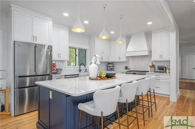 Modern kitchen with white cabinets, stainless steel refrigerator, island with white countertop, three beige bar stools, pendant lights, and wooden flooring.