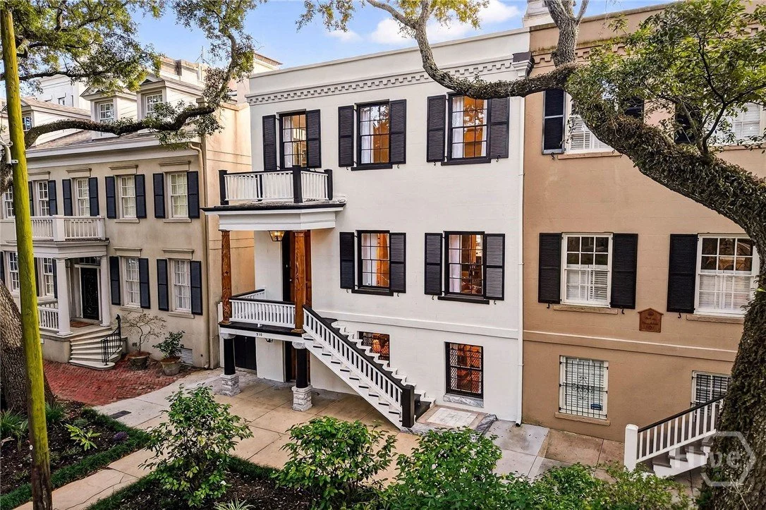 Front view of a three-story white house with black window shutters, a small balcony on the second floor, and an external staircase leading to the entrance. Surrounding trees and some plants are visible in the garden.