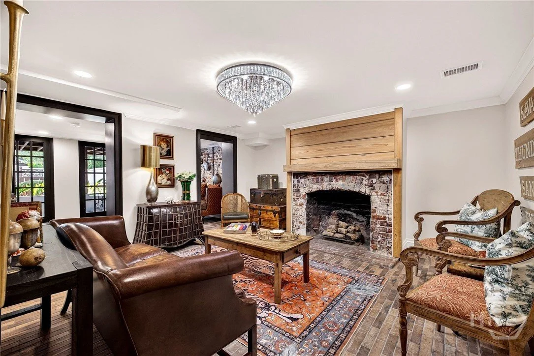 Living room with leather sofa, wooden armchairs with patterned cushions, fireplace with brick and wooden mantel, chandelier, rug, sideboard with vases and lamps, decorative wall signs, and windows with black frames.