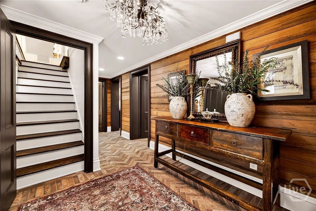 Interior view of a home entryway with wooden walls, a rustic wooden console table with two large decorative vases and greenery, framed art and a mirror, a chandelier, and a staircase leading up to another floor.