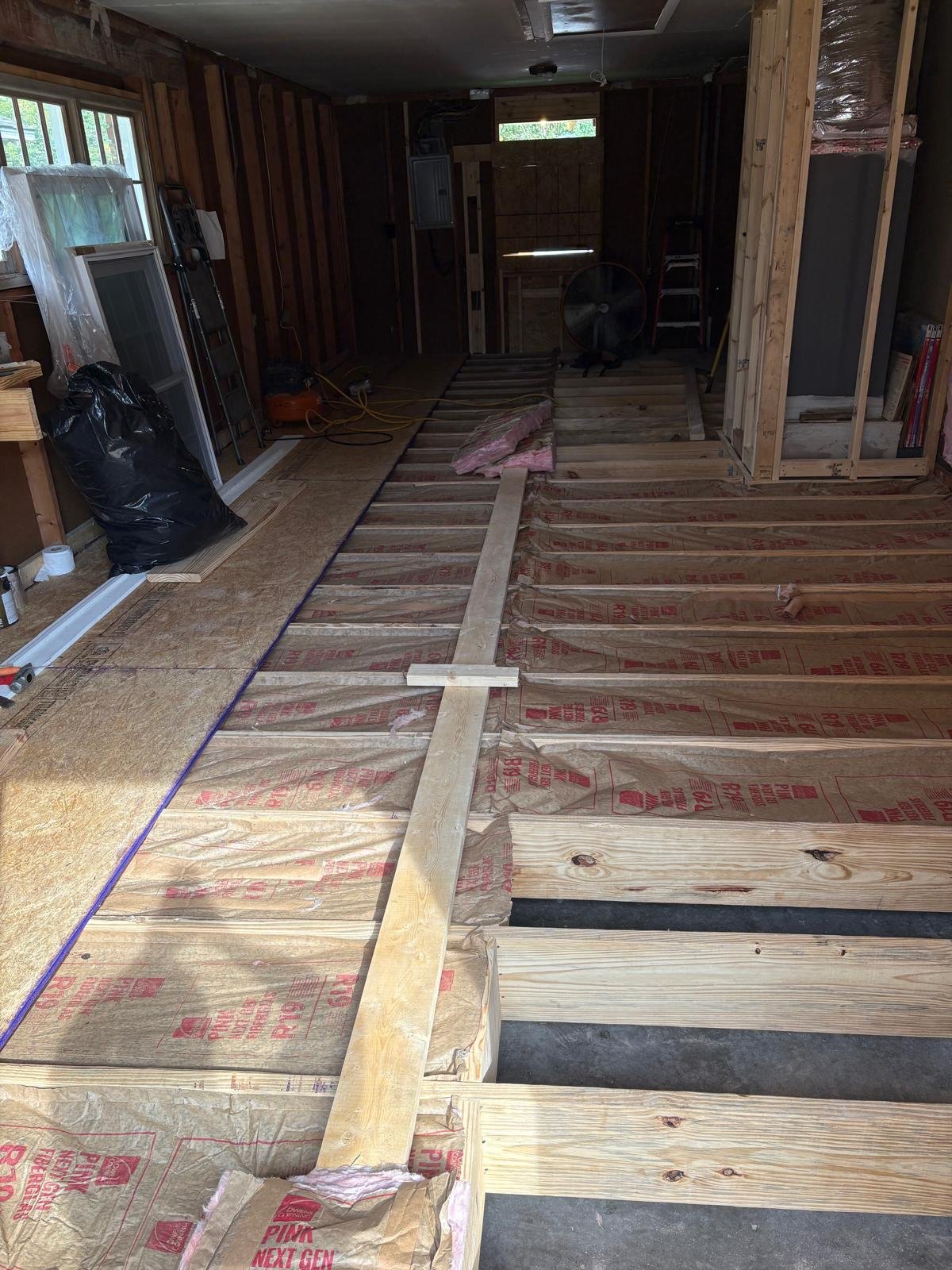 Interior of a room under renovation with exposed wooden floor joists, construction materials, a window, and tools.