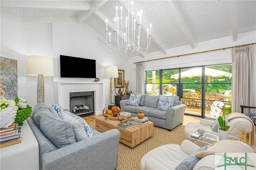 Living room with white vaulted ceiling, chandelier, gray sofas, beige armchair, wooden coffee table, fireplace, flat-screen TV, large sliding glass door with beige curtains, outdoor patio with table and umbrellas