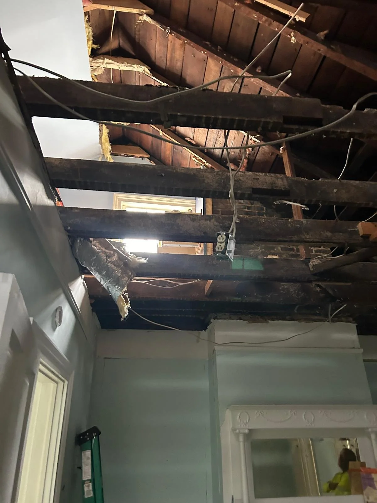 Interior of a house with a partially exposed ceiling showing wooden beams and electrical wiring, where drywall has been removed.