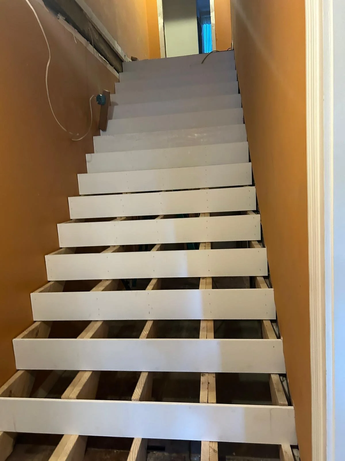 Interior staircase under construction with wooden support beams and white-painted treads, walls painted in orange, and visible wiring.