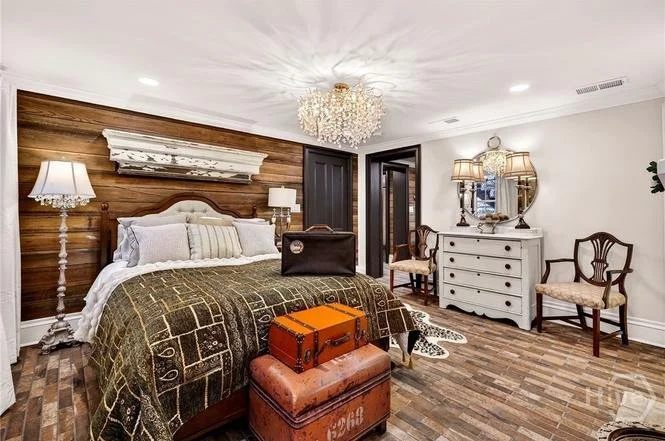 A bedroom with a large bed, wooden accent wall, chandelier, white dresser with mirror, and vintage-style chairs.