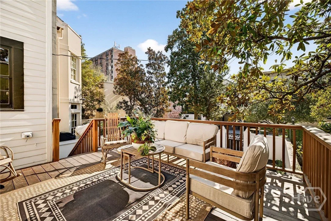 Outdoor balcony with wooden flooring and furniture including a sofa, armchairs, a coffee table, and a potted plant, surrounded by trees and buildings.