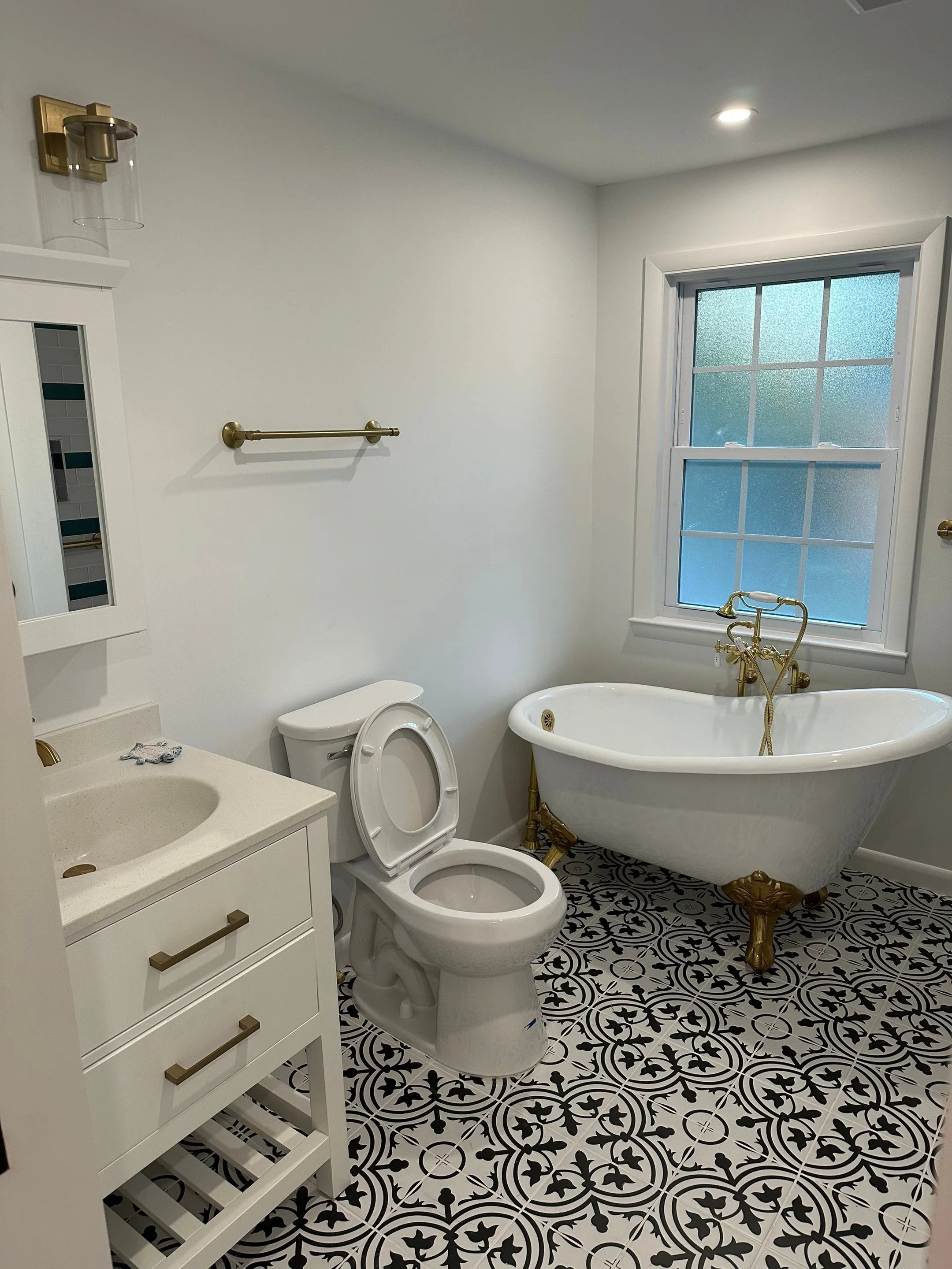 A bathroom with a vintage clawfoot bathtub with gold accents, a white toilet, a white vanity with a gold handle, a black and white patterned floor, a window with frosted glass, and gold fixtures including a towel bar and a wall sconce.