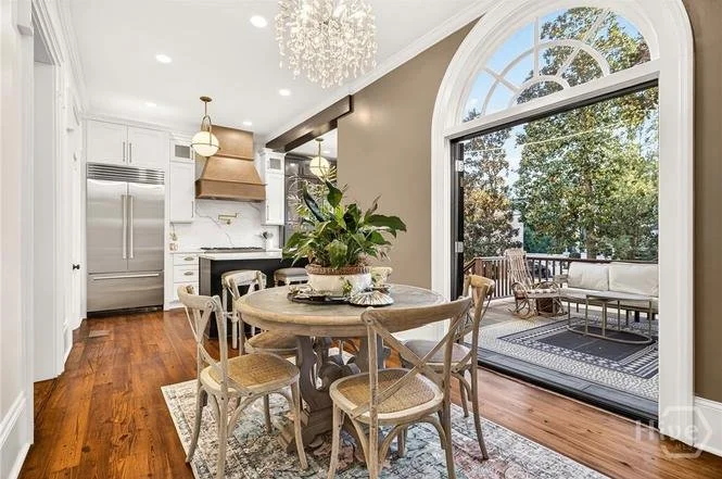 Open-concept dining area and kitchen with a large arched window leading to a balcony, wooden flooring, white cabinets, stainless steel appliances, a chandelier, and a round dining table with six chairs.