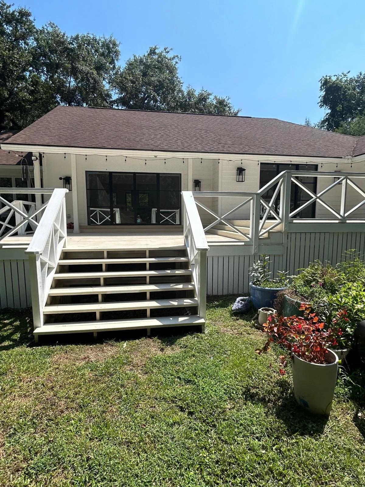 Backyard with a white wooden deck and stairs, potted plants, and string lights on a house with large windows and a brown roof.