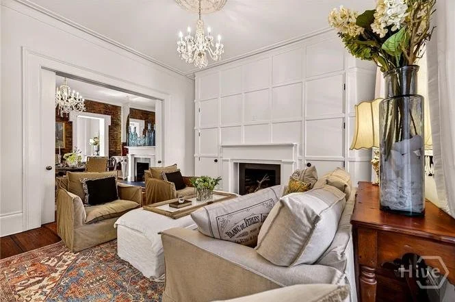 Living room with beige sofas, a white fireplace, chandelier, and a large vase of white flowers on a wooden side table.